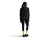 En person i Nike Dri-FIT løbejakke til kvinder (Sort/Anthracite/Reflective Silver), sorte leggings, gulgrønne sokker, sorte sneakers og solbriller står med hovedet vendt lidt til venstre.