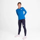 En kvinde står på en hvid baggrund iført Saysky W Clean Pace Long Sleeve, Dame, Blue med mørkeblå leggings og pink sportssko. Hun har hænderne bag ryggen og ser lige frem med et neutralt udtryk.