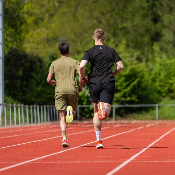 To personer løber side om side på en udendørs bane, begge i sportstøj og sneakers. Den ene har sort tøj på, mens den anden har en LiiteGuard Ground-Tech T-shirt til mænd i støvet grøn. I baggrunden ses grønne træer og hegn.