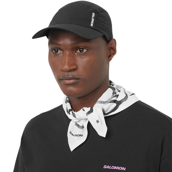 En person bærer en Salomon Shakeout Cap, Unisex, Deep Black, en sort skjorte med et lille lilla Salomon-logo og en hvid bandana med sorte mønstre om halsen. Denne åndbare hovedbeklædning skiller sig ud fra den ensfarvede hvide baggrund.