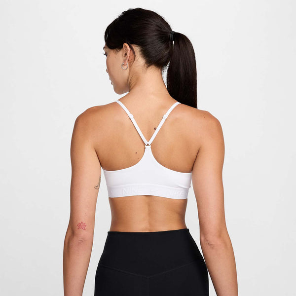 En person med langt mørkt hår i hestehale ses bagfra iført Nike Padded Adjustable Sports Bra til kvinder i White/Stone Mauve og sorte leggings. En lille rød tatovering ses på venstre arm mod en almindelig hvid baggrund.