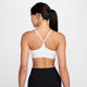 En person med langt mørkt hår i hestehale ses bagfra iført Nike Padded Adjustable Sports Bra til kvinder i White/Stone Mauve og sorte leggings. En lille rød tatovering ses på venstre arm mod en almindelig hvid baggrund.