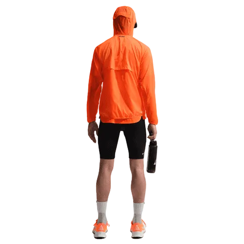 En person står med front mod en almindelig hvid baggrund iført en lys orange jakke med hætte, Nike ACG Lavaloops 1/2 Tights til mænd i Black/Summit White, hvide sokker, orange sko og med en mørk vandflaske i højre hånd.