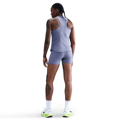 En person med flettet hår og synlige tatoveringer står med ryggen til og er iført en Nike Dri-FIT Running Tank Top i World Indigo/Reflective Silv, matchende shorts og hvide sneakers med neongrønne såler.