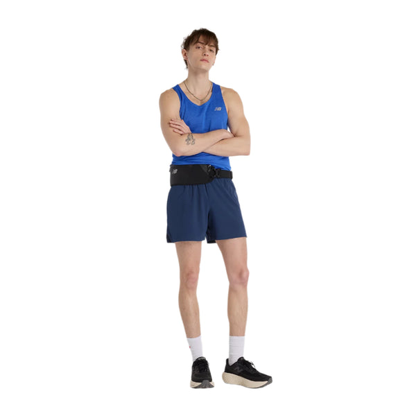 En mand står på en hvid baggrund iført en blå Athletics Tank, Herre, Blue Gemstone Heather fra New Balance, marineblå shorts, en New Balance-bæltetaske, hvide sokker og sorte løbesko - han modellerer et stilfuldt træningssæt herre.