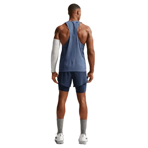 En mand kigger væk fra kameraet iført Nike AeroSwift Running Singlet, Herre, Diffused Blue/Black med matchende blå shorts, grå sokker, hvide sneakers og et hvidt kompressionsærme på højre arm mod en almindelig baggrund.