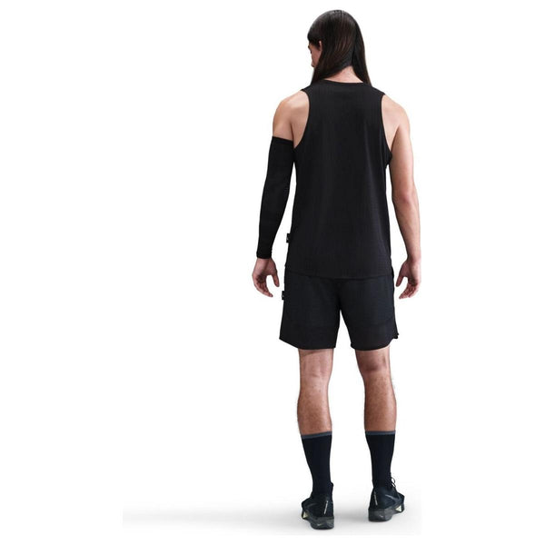 En person med langt mørkt hår kigger væk og er iført en Nike Dri-FIT Running Tank Top, Herre, Black/Reflective Silver, sammen med sorte shorts, sokker, sko og et sort ærme på venstre arm mod en almindelig hvid baggrund.