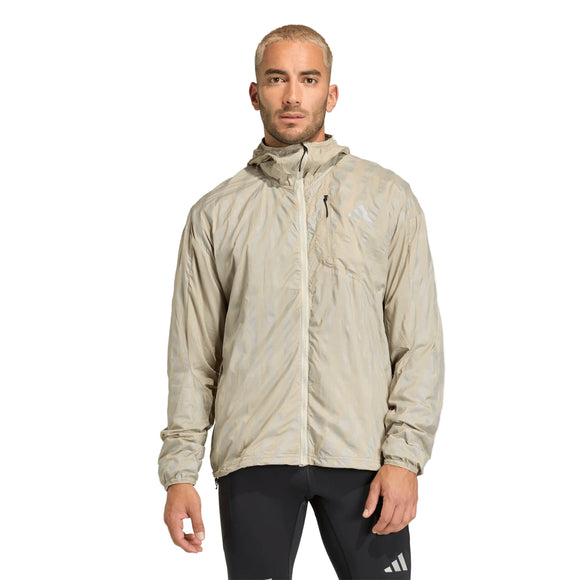 En person iført Adidas Adi365 High-Vis Jacket, Herre, Woca i lys beige med hætte og lynlås foran sammen med sorte sportsbukser med hvidt Adidas-logo står på en almindelig hvid baggrund.