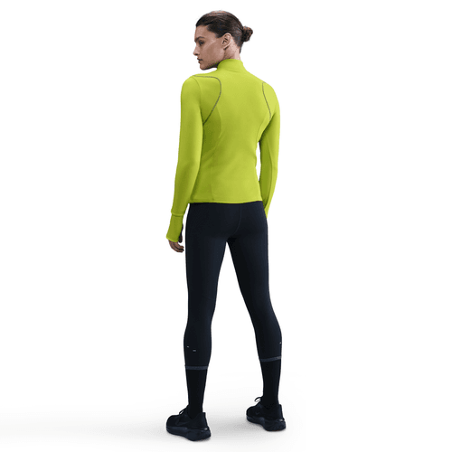 En kvinde kigger væk iført en lysegrøn Nike Therma-FIT ADV 1/2-Zip Reflective Running Top (Cyber/Reflective Silv) med sorte, tætsiddende leggings og sneakers på en almindelig hvid baggrund.
