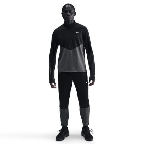 En person kigger fremad i en Nike Pacer Winterized 1/2-Zip Running Top (Herre, Anthracite/Black/Reflective Silv) med matchende sorte bukser og sneakers på en almindelig hvid baggrund.