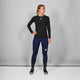 En kvinde står indendørs på en almindelig grå baggrund iført Saysky Mesh Base Layer Long Sleeve i sort, mørkeblå leggings, mønstrede sorte sokker og sorte sportssko, mens hun kigger til venstre med en hånd på hoften.