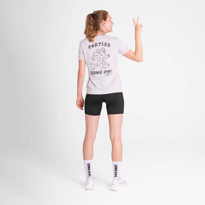 En person kigger væk og er iført Saysky W Statement Pace T-Shirt, Dame, White med en tegneserie og teksten "FARTLEK GAME ON!", sammen med sorte shorts, hvide sokker og sneakers. De holder et fredstegn op med deres højre hånd.