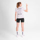 En person kigger væk og er iført Saysky W Statement Pace T-Shirt, Dame, White med en tegneserie og teksten "FARTLEK GAME ON!", sammen med sorte shorts, hvide sokker og sneakers. De holder et fredstegn op med deres højre hånd.