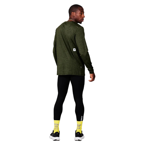 En person står med front mod kameraet iført en Saysky Clean Pace Long Sleeve, Herre, Green, sorte leggings, hvide og sorte sneakers og gule sokker med tekst på foran en almindelig lysegrå væg.