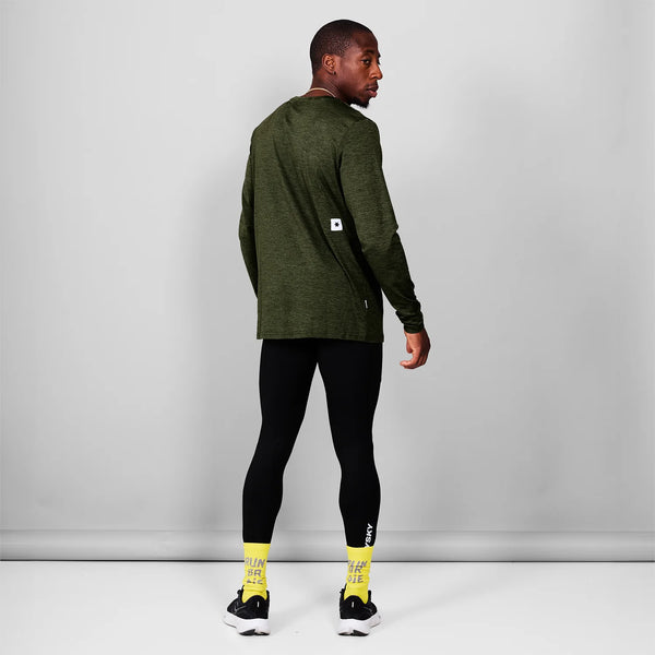 En person står med front mod kameraet iført en Saysky Clean Pace Long Sleeve, Herre, Green, sorte leggings, hvide og sorte sneakers og gule sokker med tekst på foran en almindelig lysegrå væg.
