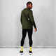 En person står med front mod kameraet iført en Saysky Clean Pace Long Sleeve, Herre, Green, sorte leggings, hvide og sorte sneakers og gule sokker med tekst på foran en almindelig lysegrå væg.