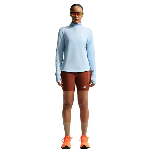 En person kigger fremad i en Nike ACG Half Zip Running Top, Dame, Hydrogen Blue/Summit White, rustne Adidas-shorts, orange løbesko, brune sokker og reflekterende sportssolbriller på en hvid baggrund.