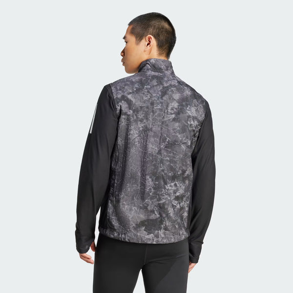 En mand kigger væk fra kameraet iført Adidas Otr Sd Vest M til mænd i Grefou/Black med ensfarvede sorte ærmer og sorte bukser på en almindelig lys baggrund.