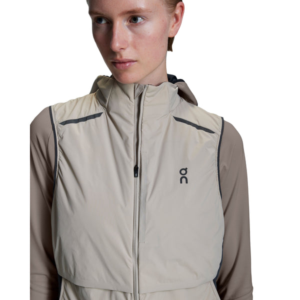 En person bærer On Weather Vest, Dame, Desert/Eclipse - en let beige vest med hætte og brystlogo - over en taupefarvet langærmet skjorte og kigger lidt til siden mod en hvid baggrund.