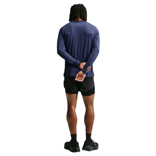 En person står med ryggen til og har en Nike Dri-FIT ADV langærmet løbetrøje til mænd i farven Midnight Navy/Reflective Silver på, kombineret med sorte shorts og sneakers. Armene er bag ryggen, og hænderne er foldet. Det avancerede Dri-FIT ADV-materiale i Nike-trøjen sikrer komfort og høj ydeevne på den ensfarvede hvide baggrund.