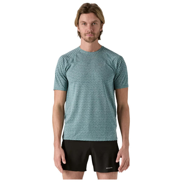 En mand står og kigger fremad iført en Patagonia M's Ridge Flow Shirt, Herre, Wetland Blue - en åndbar løbetrøje - og sorte shorts mod en almindelig hvid baggrund.