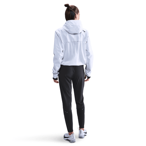 En person står med ryggen til kameraet iført en lys jakke med hætte, Nike Dri-FIT Mid-Rise løbebukser til kvinder i sort/reflekterende sølv og sportssko på en almindelig hvid baggrund.
