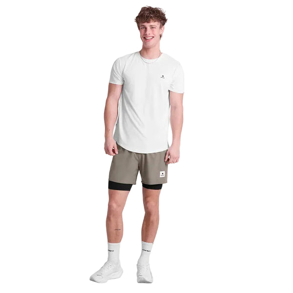 En person smiler mod en hvid baggrund iført en hvid t-shirt, Saysky Pace 2 in 1 Shorts 5'' Herre i Taupe, hvide sokker og sneakers. Med kort, krøllet hår er dette look ideelt til maratonløbere, der ønsker komfort og stil.