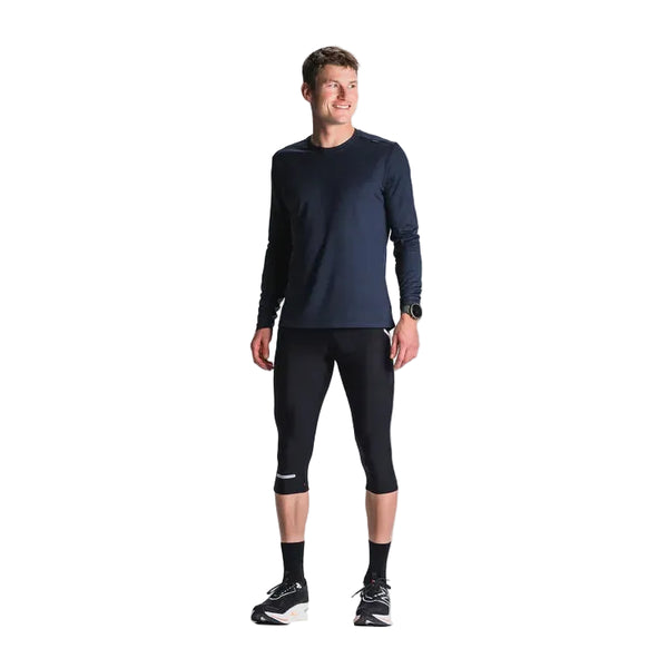 En mand står sidelæns og smiler i en mørk langærmet skjorte og Fusion 3/4 Tights Unisex SuperBlack, sammen med sorte sokker, sko og et ur på venstre håndled mod en lys baggrund.