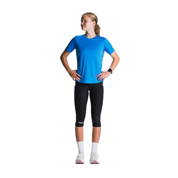 En person står lidt sidelæns med hænderne på hofterne iført blå t-shirt, Fusion 3/4 Tights i SuperBlack, hvide sokker og hvide sneakers på en almindelig lys baggrund.