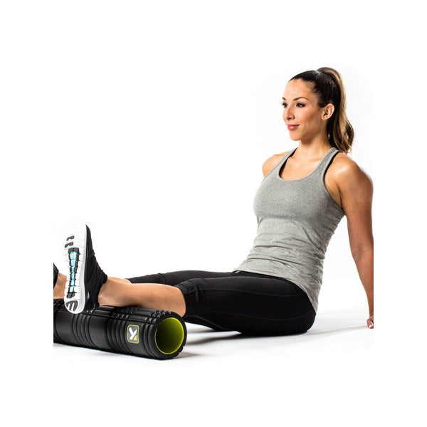 En kvinde i grå tanktop og sorte leggings bruger Trigger Point Grid 2.0 Foam Roller, Unisex, Black under sin højre læg, mens hun sidder på gulvet med hænderne bag sig som støtte og venstre knæ bøjet.