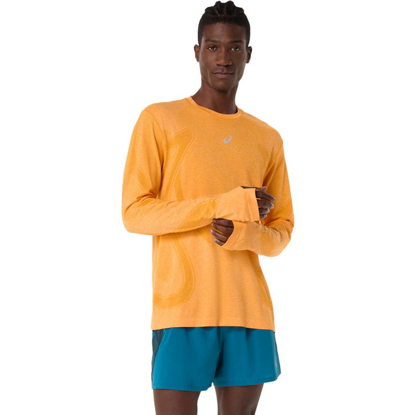 En person står med ansigtet fremad og er iført en orange Asics Road Seamless Ls Top, Herre, Yamabuki med huller til tommelfingrene og blågrønne sportsshorts på en almindelig hvid baggrund.