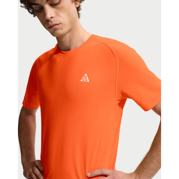 En mand står med hænderne på hofterne iført Nike ACG Dfadv Solar Chase Ss træningstop herre i Safety Orange/Summit White med et lille hvidt logo på brystet. Den ensfarvede, lyse baggrund passer til udendørs træning.
