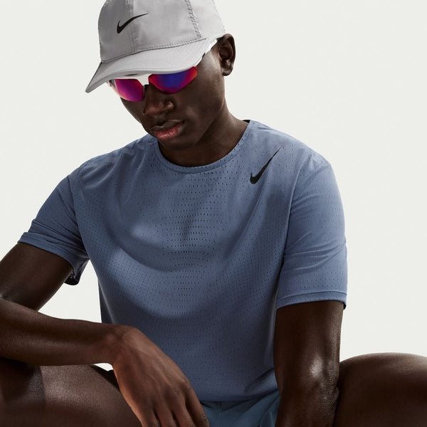 En person iført en lysegrå Nike-kasket, reflekterende solbriller med pink glas og en Nike Dfadv Aroswft Ss Top, Herre, Diffused Blue/Black lavet af svedtransporterende løbetøj sidder på en almindelig hvid baggrund.