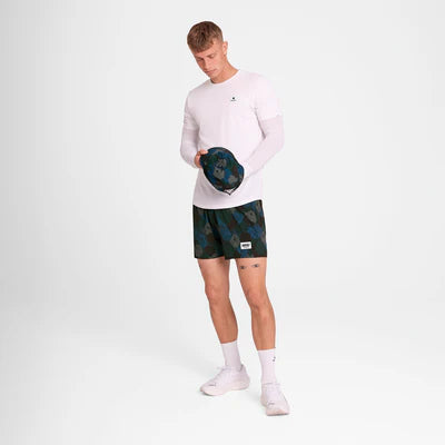 En mand står i et hvidt rum iført en langærmet hvid skjorte, Saysky Flower Pace Shorts 5" Herre in Blue Aop, hvide sokker og sneakers. Han kigger ned, mens han holder en mørk camo-baseballkasket i hænderne.