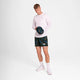 En mand står i et hvidt rum iført en langærmet hvid skjorte, Saysky Flower Pace Shorts 5" Herre in Blue Aop, hvide sokker og sneakers. Han kigger ned, mens han holder en mørk camo-baseballkasket i hænderne.