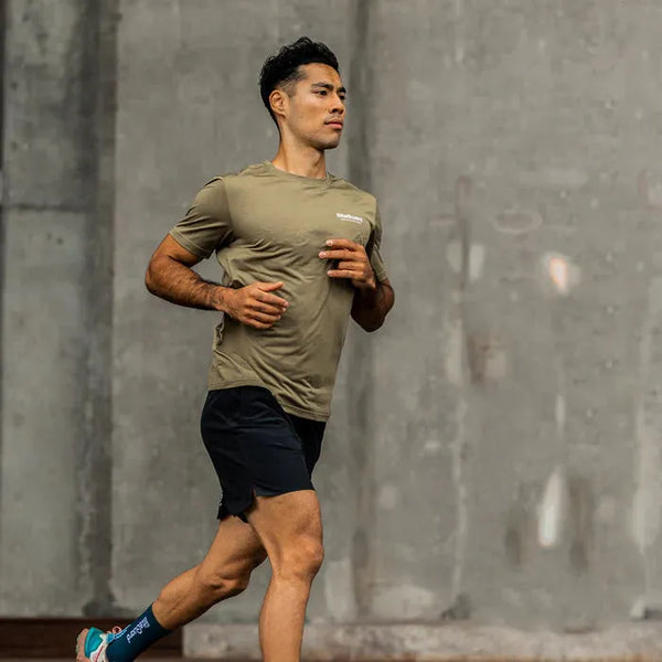 En mand iført en LiiteGuard Ground-Tech T-shirt til mænd i farven Dusty Green jogger udendørs foran en stor betonmur.