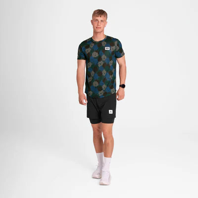 En mand med kort blondt hår og et neutralt udtryk bærer Saysky Flower Combat T-Shirt, Herre, Blue Aop, sammen med sorte shorts, hvide sokker og hvide sneakers på en almindelig hvid baggrund.