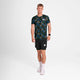 En mand med kort blondt hår og et neutralt udtryk bærer Saysky Flower Combat T-Shirt, Herre, Blue Aop, sammen med sorte shorts, hvide sokker og hvide sneakers på en almindelig hvid baggrund.