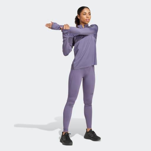 En kvinde iført en matchende lilla Adidas Otr B Hzip træningstrøje dame med halv lynlås og sorte sneakers strækker sin højre arm over brystet mod en almindelig lysegrå baggrund.