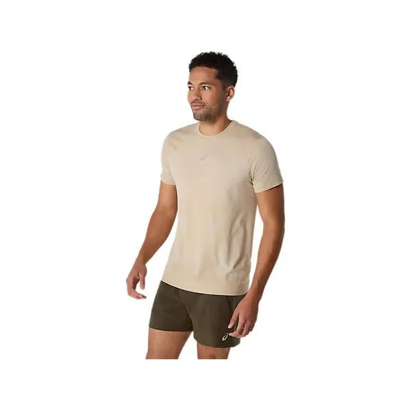 En mand står på en hvid baggrund iført en Asics Road Seamless SS Top, Herre, Cream. Han ser lidt til venstre med afslappede arme, hvilket fremhæver den åndbare åndbar træningstrøje, der er designet til hverdagskomfort.