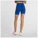 En kvinde står med armene over kors og viser New Balance Sleek Pocket High Rise Short 6" i Galaxy Blue og hvide sneakers, set bagfra mod en almindelig hvid baggrund.