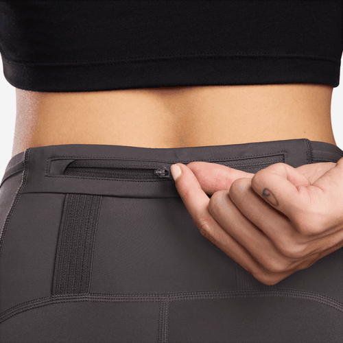 En person iført Nike Trail 7/8 Tights, Dame, i Thunder Grey/Black åbner en lille lynlåslomme i den bagerste linning, så kun underkroppen og hånden er synlig.