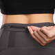 En person iført Nike Trail 7/8 Tights, Dame, i Thunder Grey/Black åbner en lille lynlåslomme i den bagerste linning, så kun underkroppen og hånden er synlig.