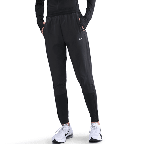 En kvinde iført sorte Nike Dri-FIT Mid-Rise Running Pants (Black/Reflective Silver), en sort langærmet top og hvide Nike-sneakers står med hænderne i lommerne mod en hvid baggrund. Perfekt træningstøj dame-stil.