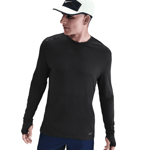 En mand i Nike Dri-FIT ADV Wool Long-Sleeve Running Top for men i Black/Anthracite, en hvid og sort Nike-kasket og blå shorts står let foroverbøjet med venstre tommelfinger gennem et hul i ærmet.