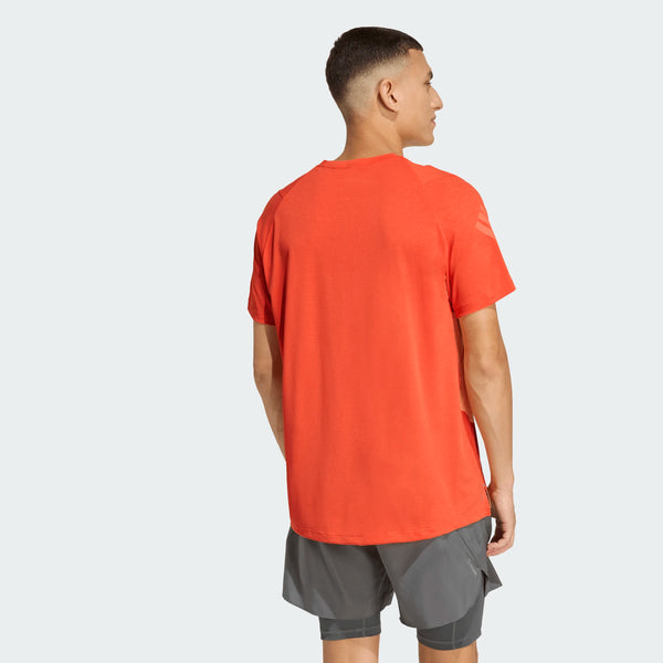 En person bærer den knaldorange Adi365 Running Tee, Herre, Duor fra Adidas med grå shorts og kigger væk for at vise bagsiden af denne adidas løbetrøje - ideel til løbetræning - mod en almindelig lys baggrund.
