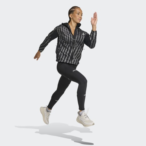 En kvinde bærer Adidas Adi365 Jacket, Dame, Black/Refsil med sorte leggings og hvide sportssko, mens hun poserer i luften på en almindelig lys baggrund.