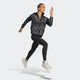 En kvinde bærer Adidas Adi365 Jacket, Dame, Black/Refsil med sorte leggings og hvide sportssko, mens hun poserer i luften på en almindelig lys baggrund.