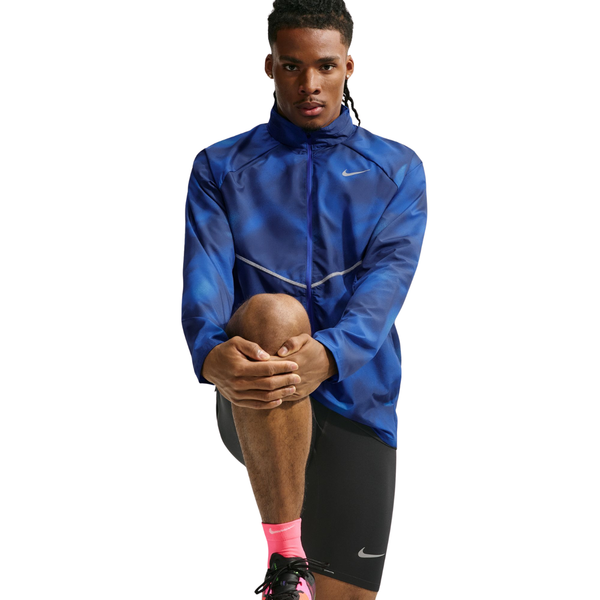 En person iført Nike Miler UV Repel-løbejakken til mænd i farvekombinationen Midnight Navy/Reflective Silver, sorte Nike-shorts og lyserøde sokker strækker sig ved at løfte det ene knæ op. Baggrunden er ensfarvet hvid.