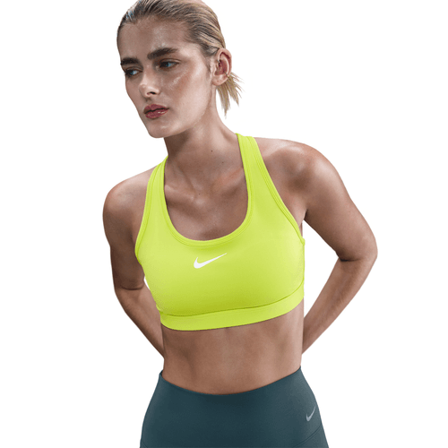 En kvinde med tilbagestrøget blondt hår bærer Nike Swoosh Medium Sports Bra til kvinder i Cyber/White og mørke leggings, mens hun står med hænderne bag ryggen og kigger lidt til siden mod en hvid baggrund.